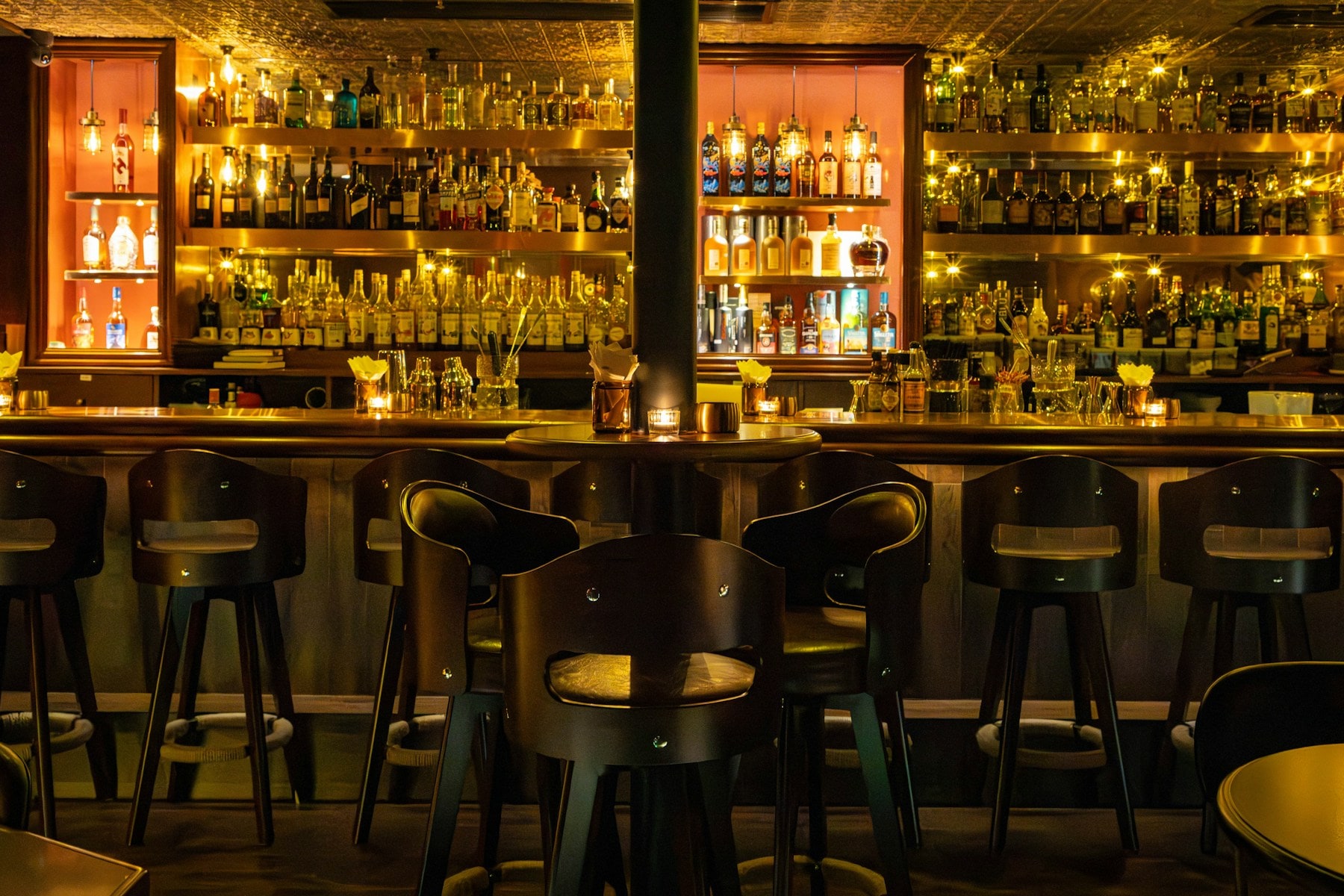 A warmly lit back bar stacked with bottles above a row of stools.